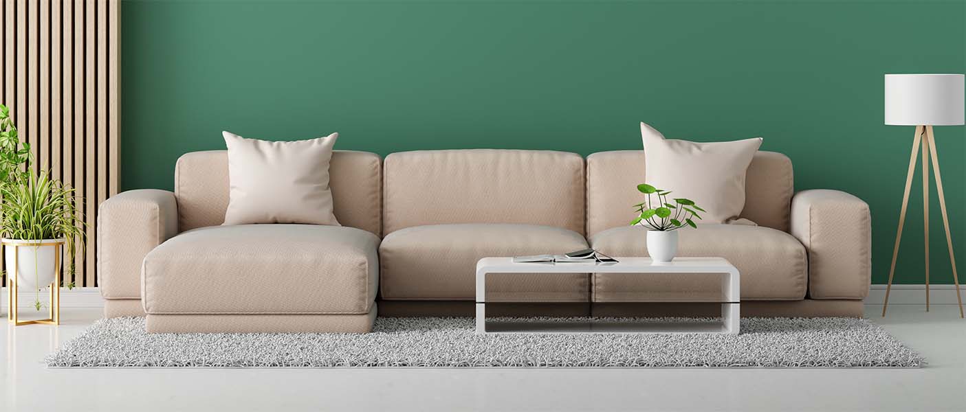 Beige Wohnlandschaft Sofa mit Kissen auf modernem Teppich, angrenzend an einen Couchtisch mit Pflanze. Grünes Wandpanel und dekorative Lampe im Hintergrund. Modernes Wohnzimmermöbel-Design, gemütliches Ambiente, stilvolle Inneneinrichtung.